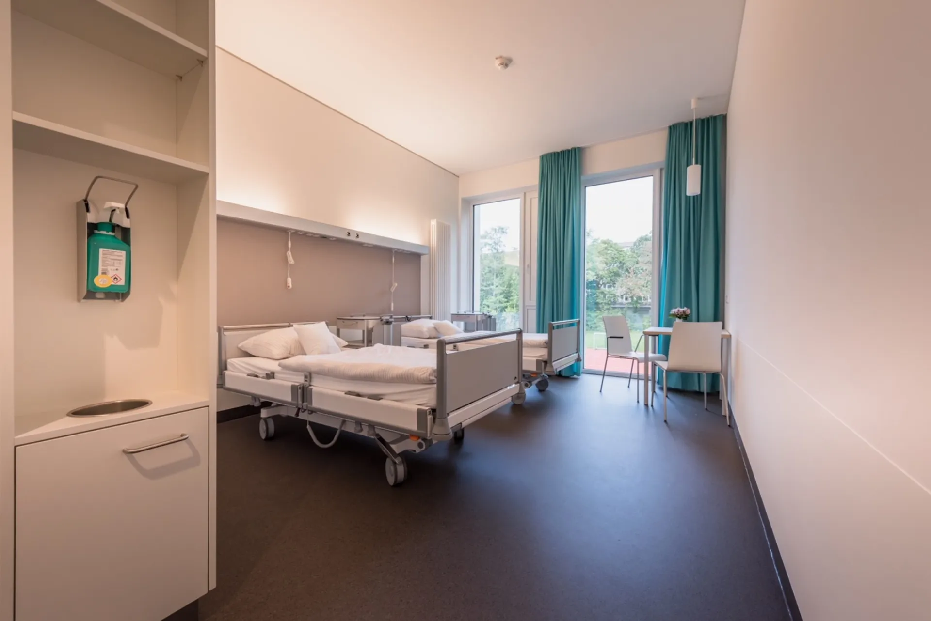 Hospital room with two beds, chairs and a large window with green curtains
